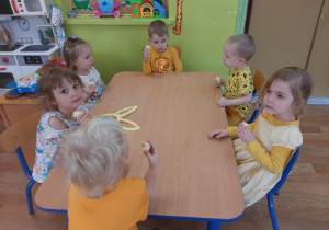 Dzieci ubrane na żółto siedzą przy stoliku i jedzą banany